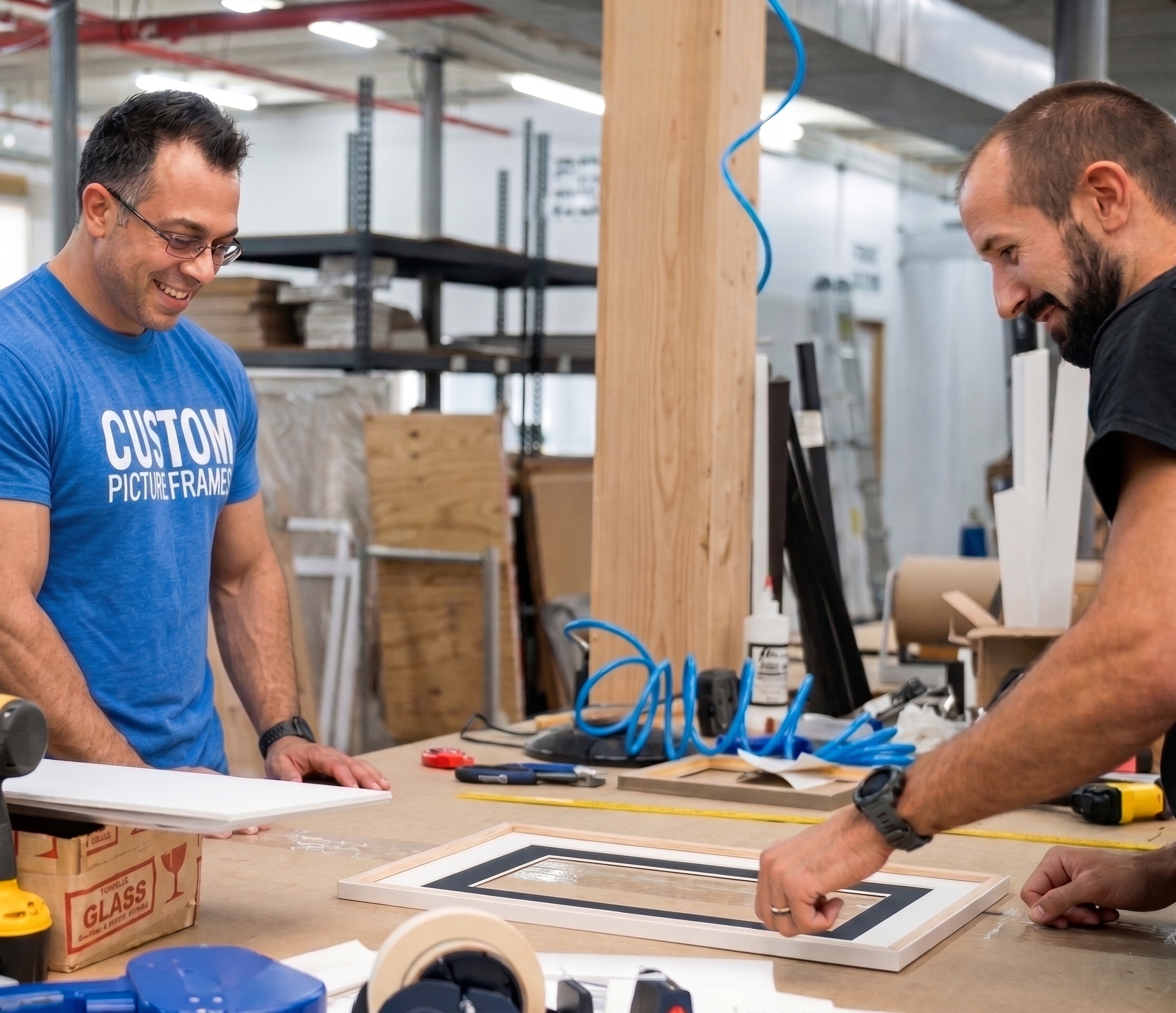Two framers smiling at the workbench in the Somerset, NJ workshop while assembling a matted frame, with a Custom Picture Frames branded shirt visible